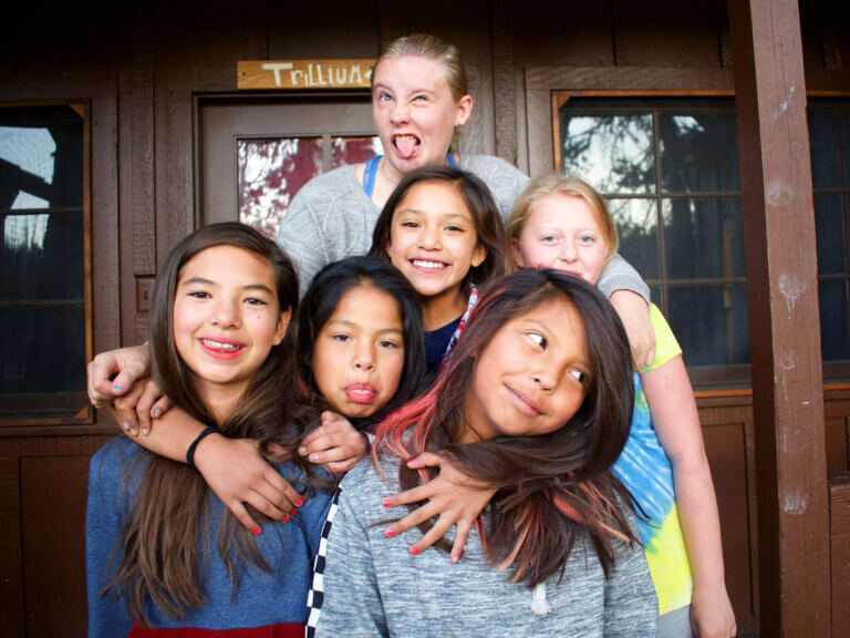 A group of girls pose with silly smiles in front of their cabin.